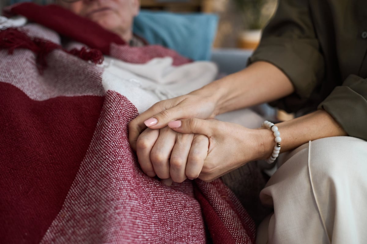 Mano de una persona sosteniendo con cuidado la mano de un adulto mayor bajo una manta